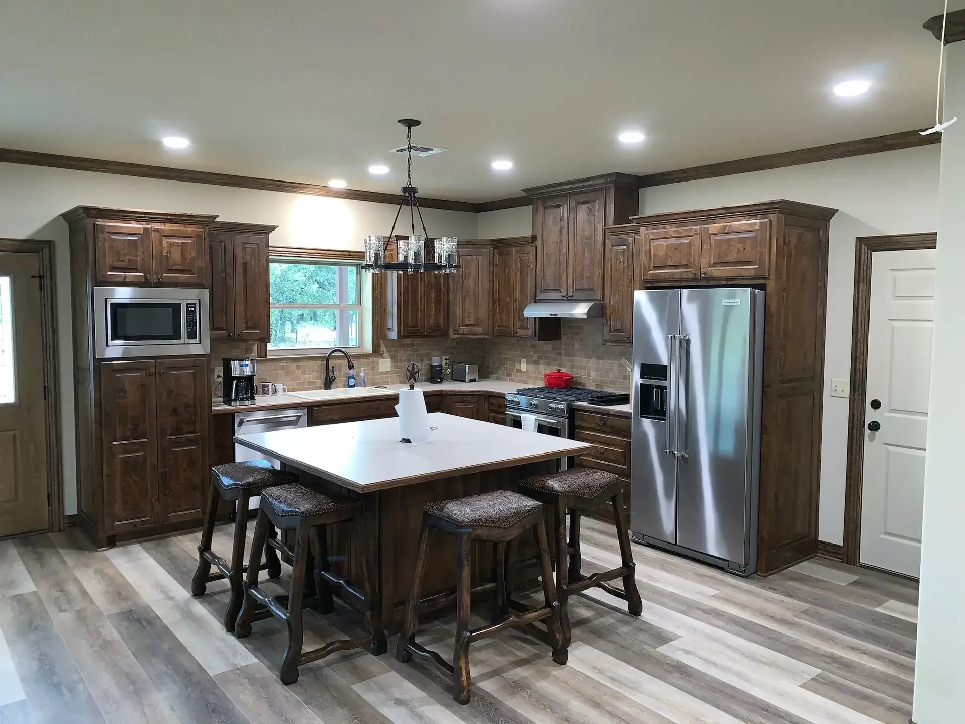 Shows the inside of a house, there is gray wood-like linoleum flooring, dark brown cabinets and a matching island. There are dark brown wooden barstools with worked leather seats.